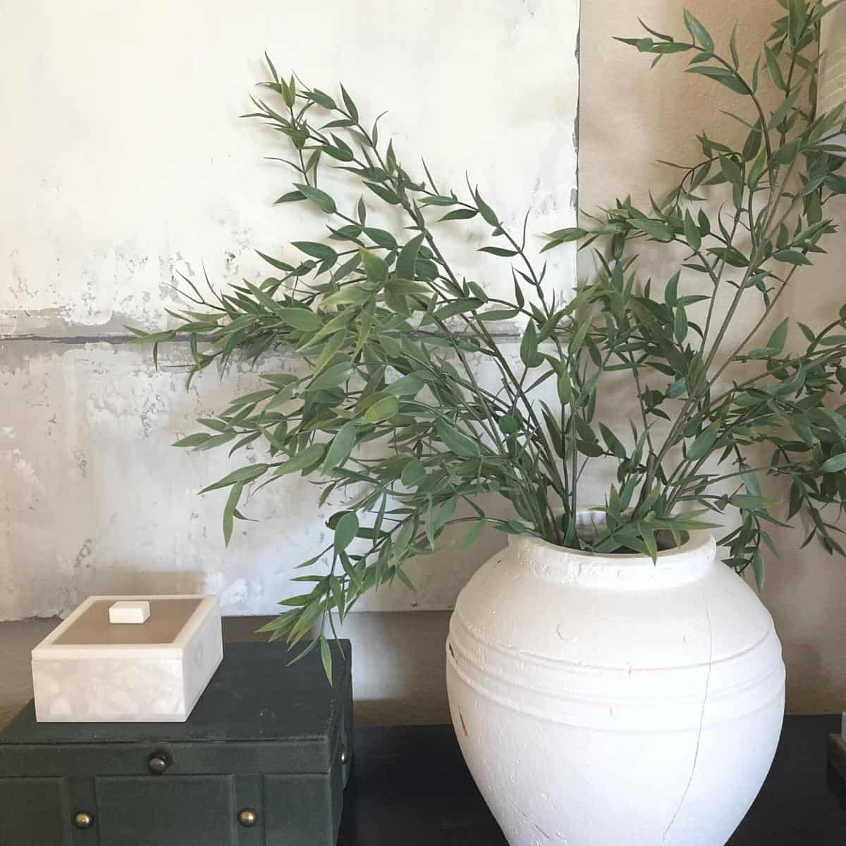 Abstract wall art behind a white vase with large, faux stems, styled beside a large green felt box with a smaller, white alabaster box on top.