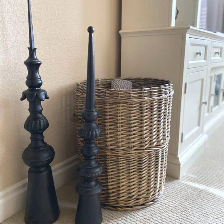 Antique architectural salvage French black finials stand in front of a tall, large hand-woven antique basket with leather handles in a cognac finish. An off-white entertainment center is to the side in the back.