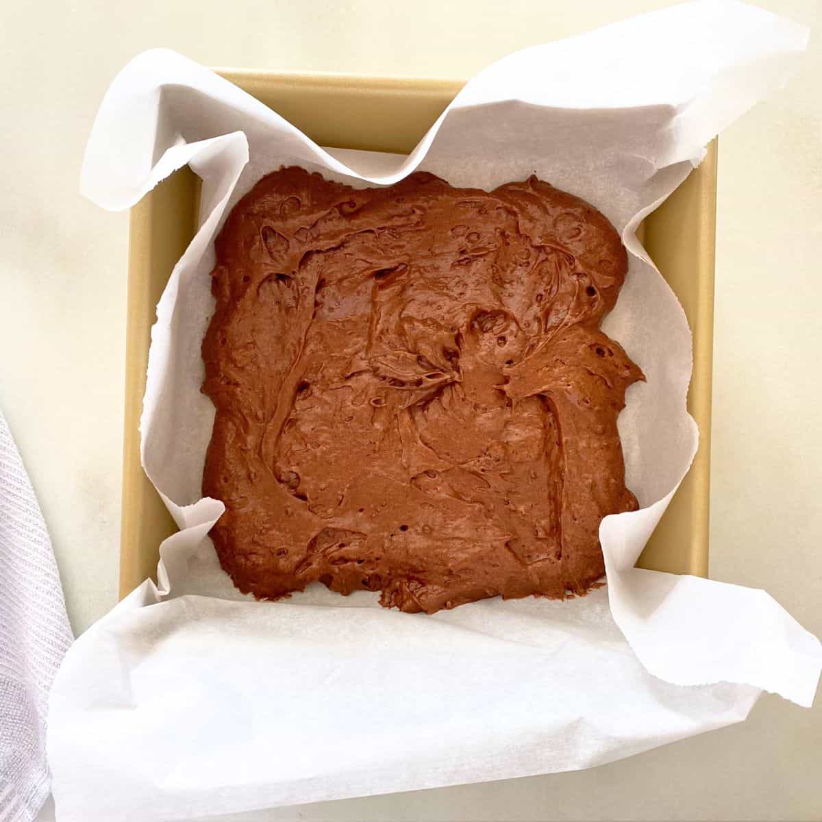 Uncooked brownie batter in a gold baking pan lined with parchment paper