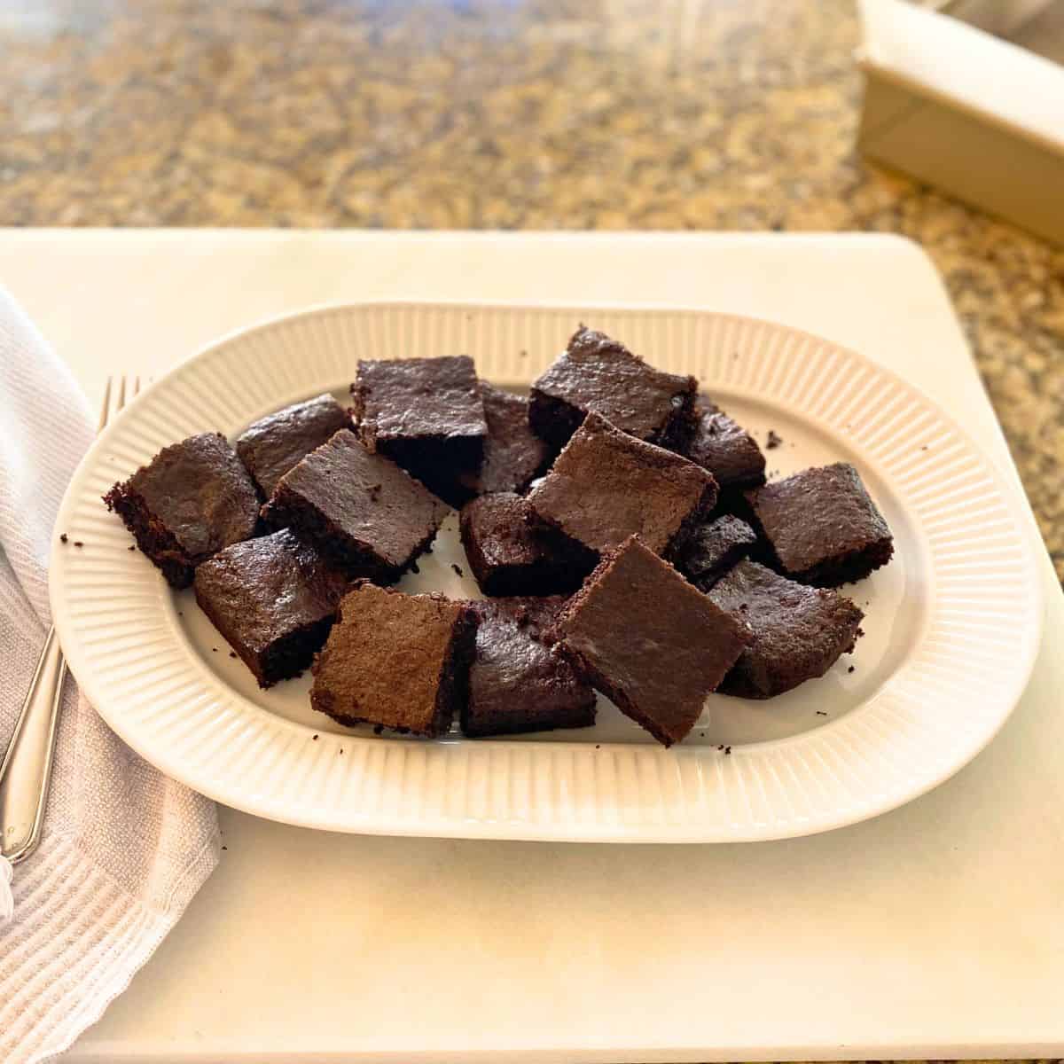 Baked brownies cut into squares stacked on a fluted white porcelain serving platter with a fork next to them