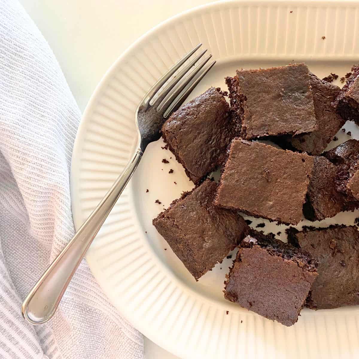 Baked brownies cut into squares stacked on a fluted white porcelain serving platter with a fork next to them