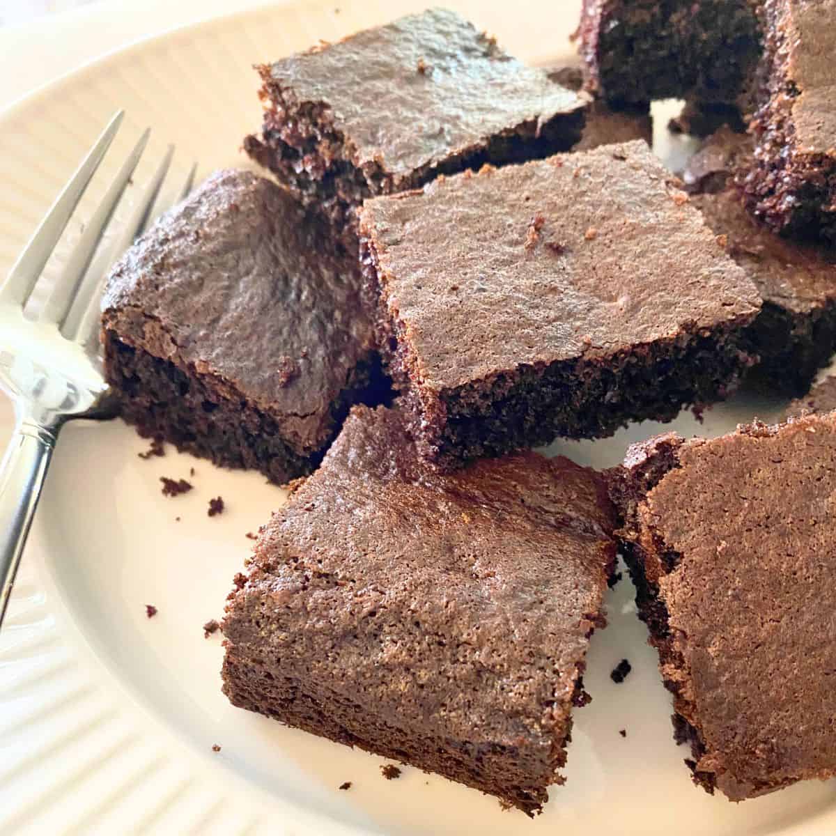 Baked brownies cut into squares stacked on a fluted white porcelain serving platter with a fork next to them