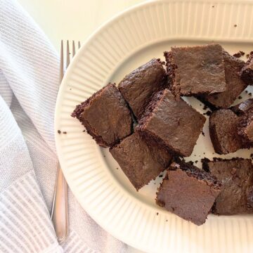 Baked brownies cut into squares stacked on a fluted white porcelain serving platter with a fork next to them