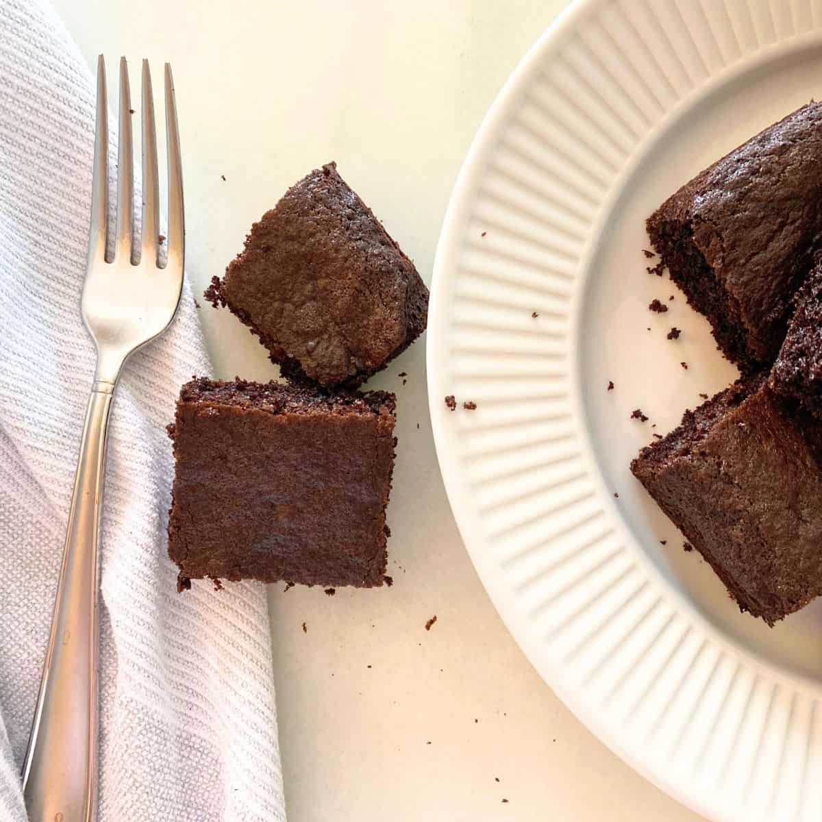 Baked brownies cut into squares stacked on a white marble pastry board next to a silver fork