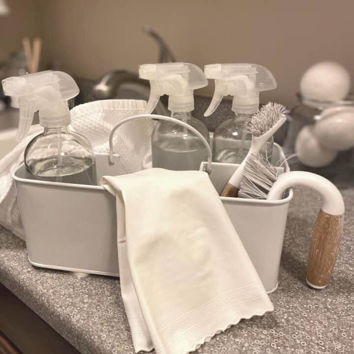 White metal cleaning caddy with cleaning products inside, including three glass spray bottles, white cleaning gloves, a scrub brush and cotton cloth.