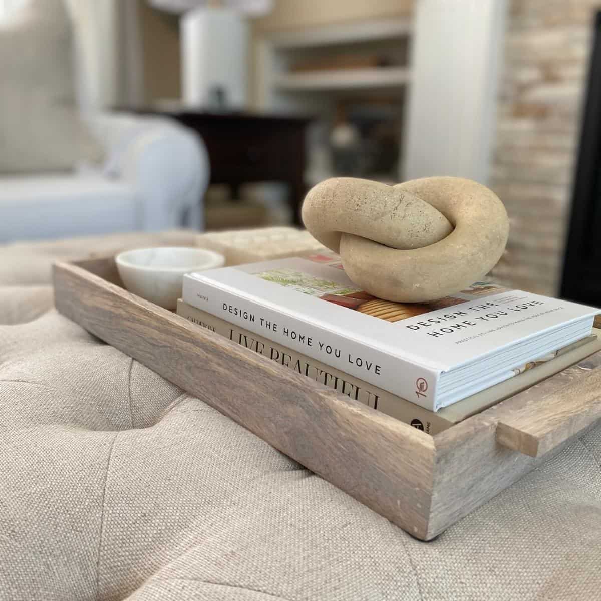 Twist decor object atop a stack of coffee table books styled in a light wood tray, beside a small marble bowl and bone box.