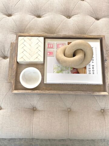 Twist decor object atop a stack of coffee table books styled in a light wood tray, beside a small marble bowl and bone box.
