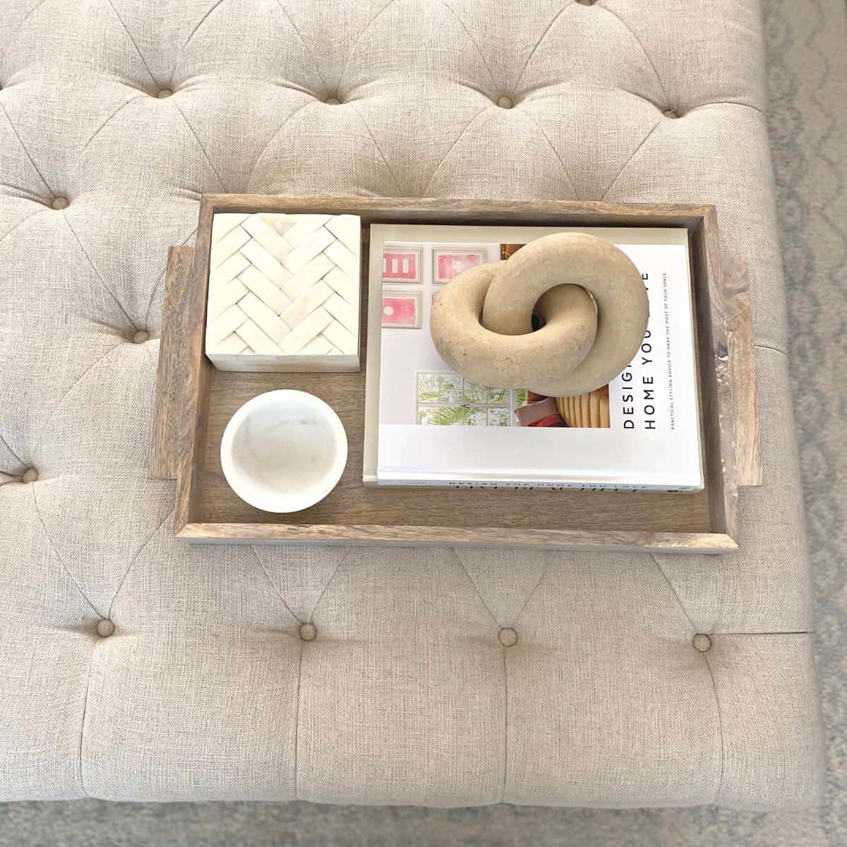 Twist decor object atop a stack of coffee table books styled in a light wood tray, beside a small marble bowl and bone box.