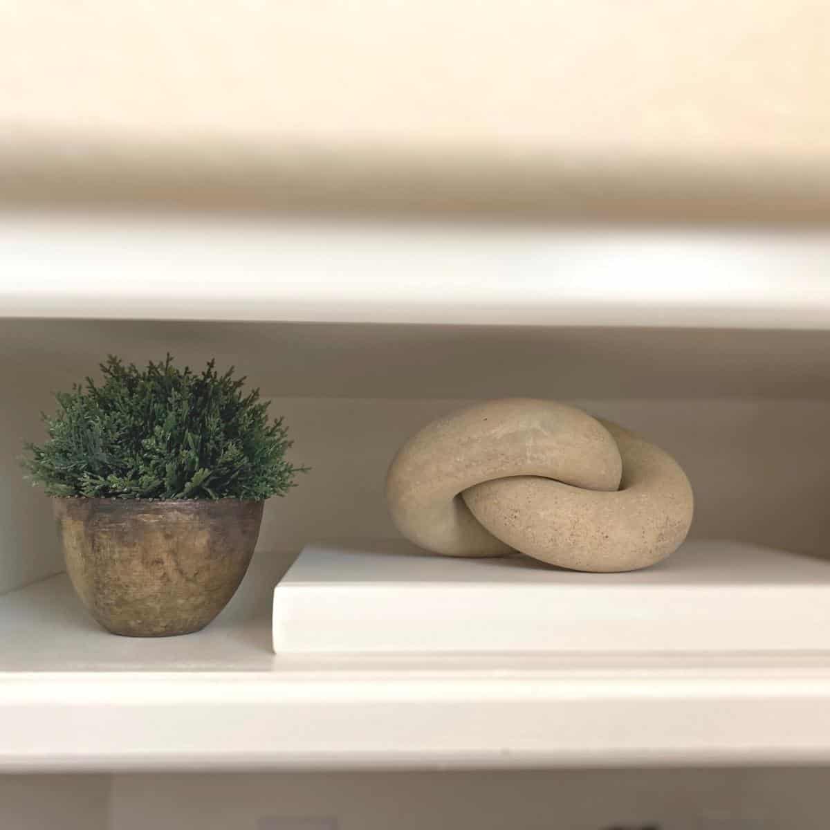 Shelf styling of a small faux plant in brass container next to white book with a twist object sitting on top.