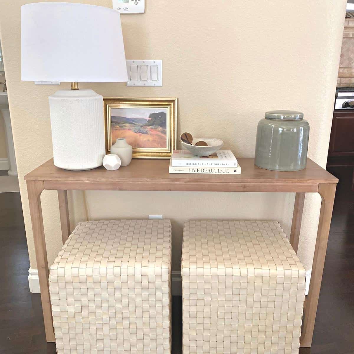 Entryway console table and two square ottomans styled with white lamp, small objects, a stack of two books and a large green vase. A small art piece stands behind.