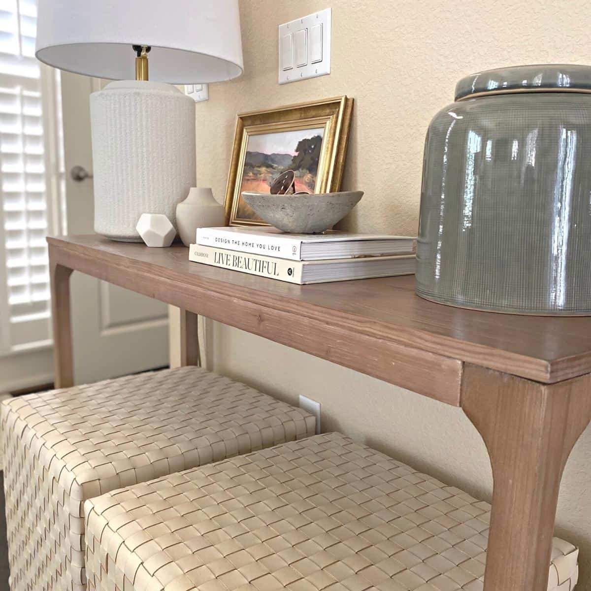 Entryway console table and two square ottomans styled with white lamp, small objects, a stack of two books and a large green vase. A small art piece stands behind.