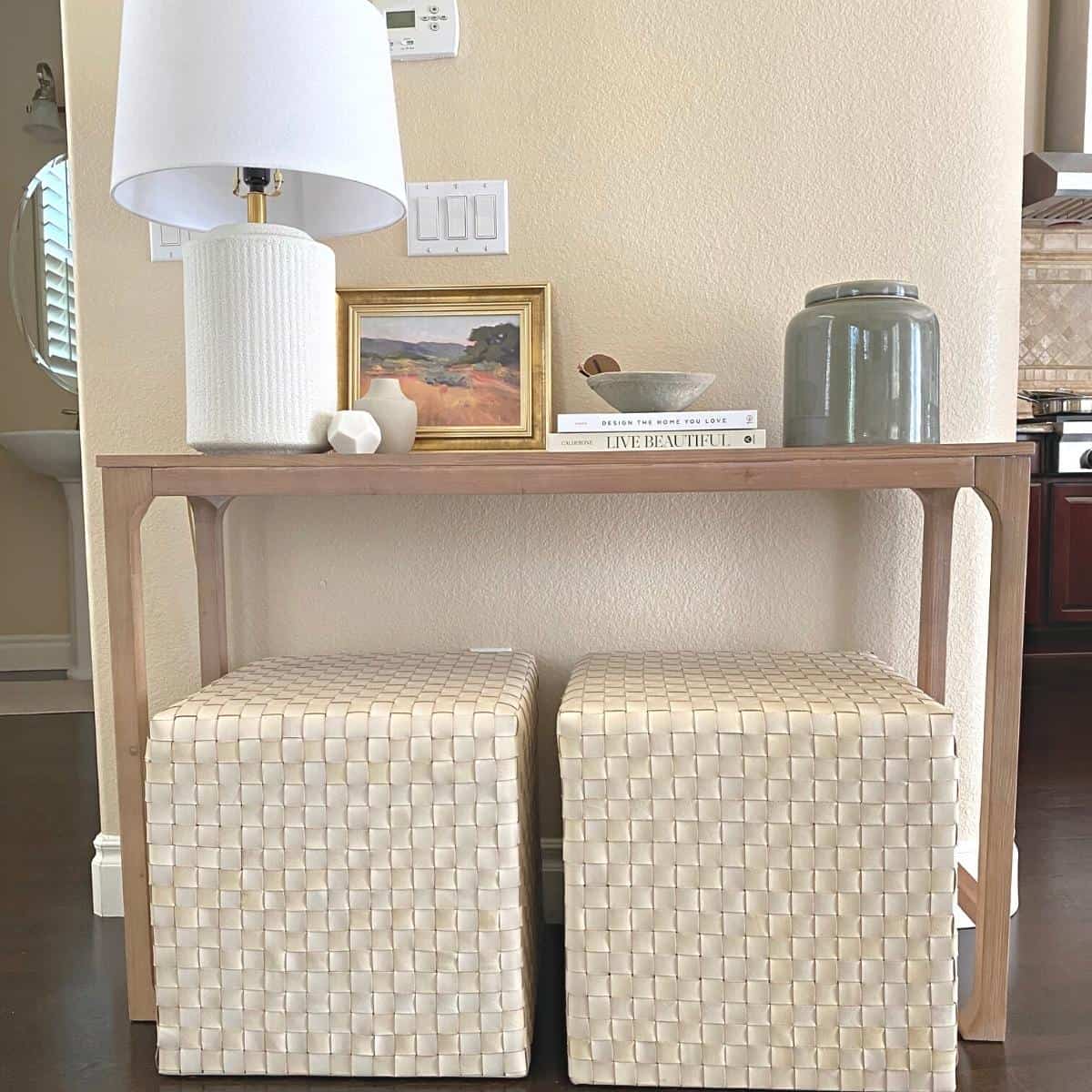 Entryway console table and two square ottomans styled with white lamp, small objects, a stack of two books and a large green vase. A small art piece stands behind.