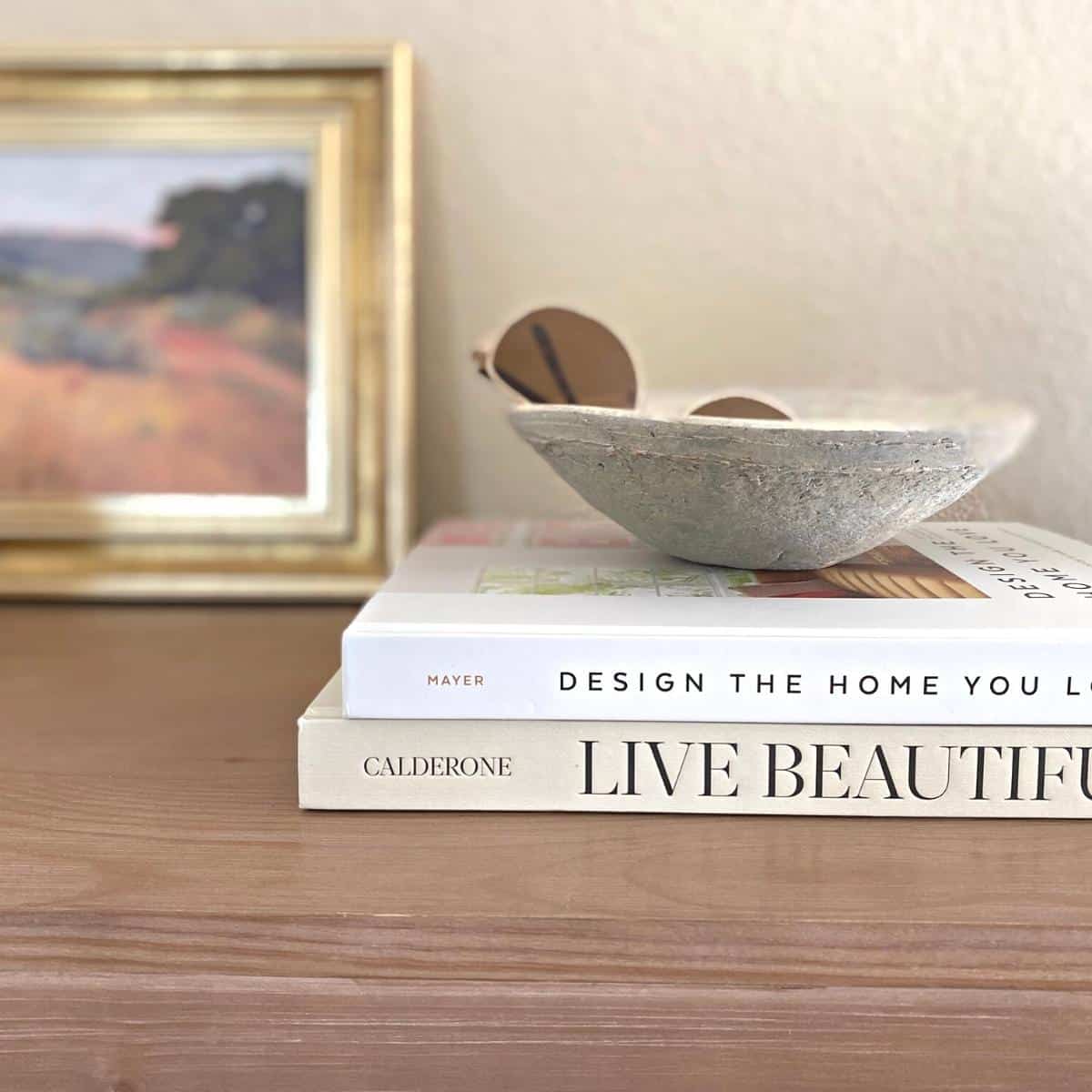 Small gold framed artwork styled next to a stack of two neutral books that has a small grey textured bowl containing sunglasses on top.