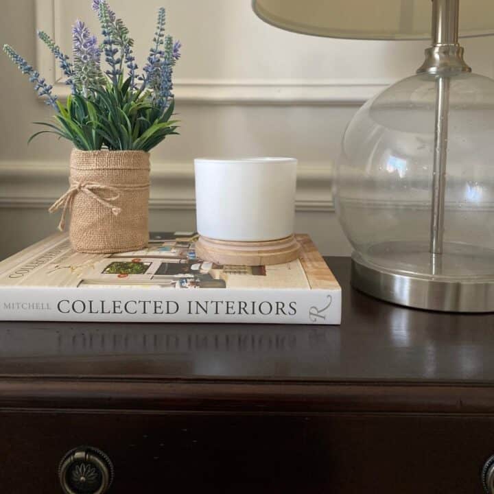 A dark wood nightstand with a glass lamp, white candle, faux lavender in a burlap sack greenery, and the book, Collected Interiors.