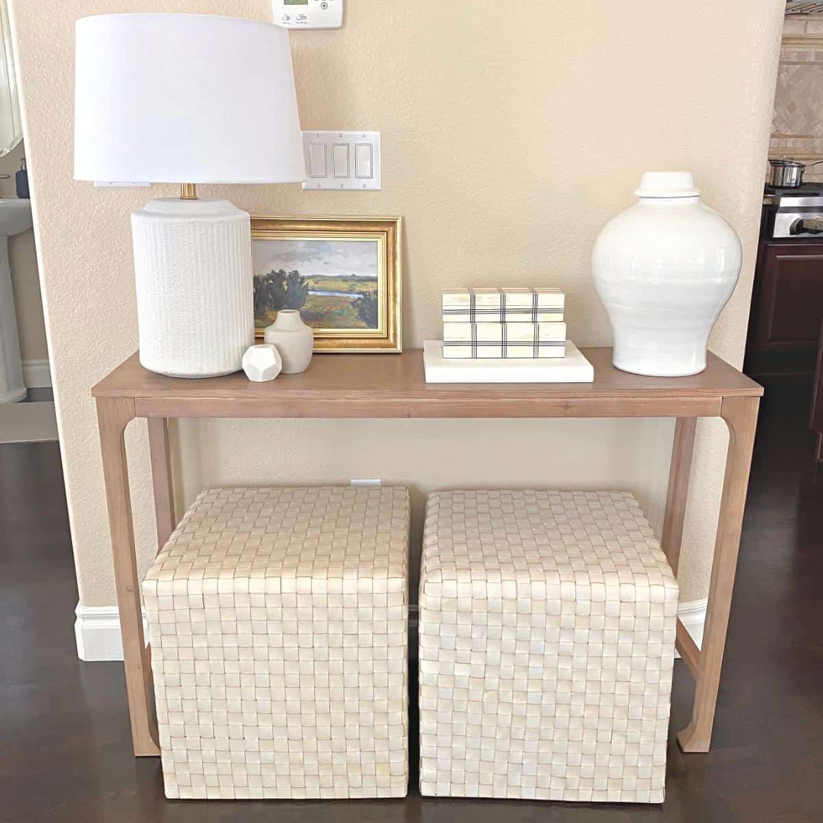 Console table styled with a white textured lamp on the left, small framed art next to a set of stacked wide grid bone decorative boxes on a white book in the middle and a large white vase on the right