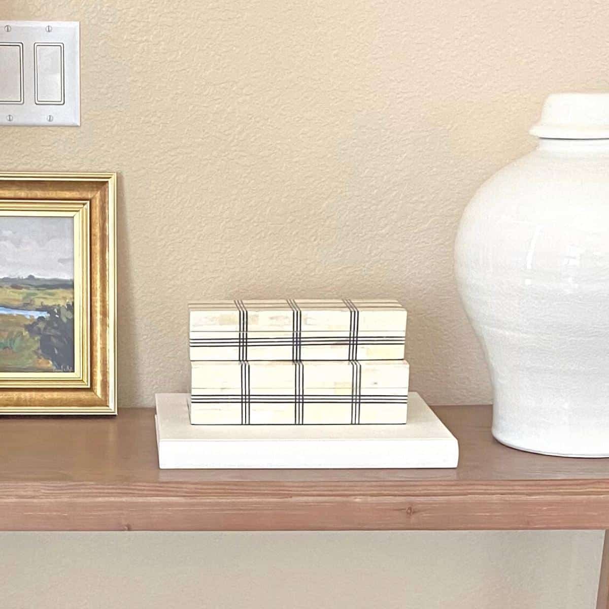 Wide grid box boxes atop a white book, flanked by small gold framed art on the left and a large white vase on the right