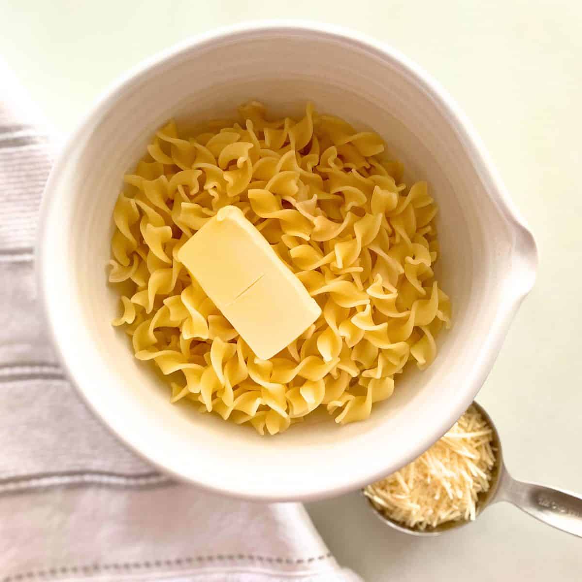 Cooked noodles topped with butter in white clay mixing bowl, parmesan sits beside