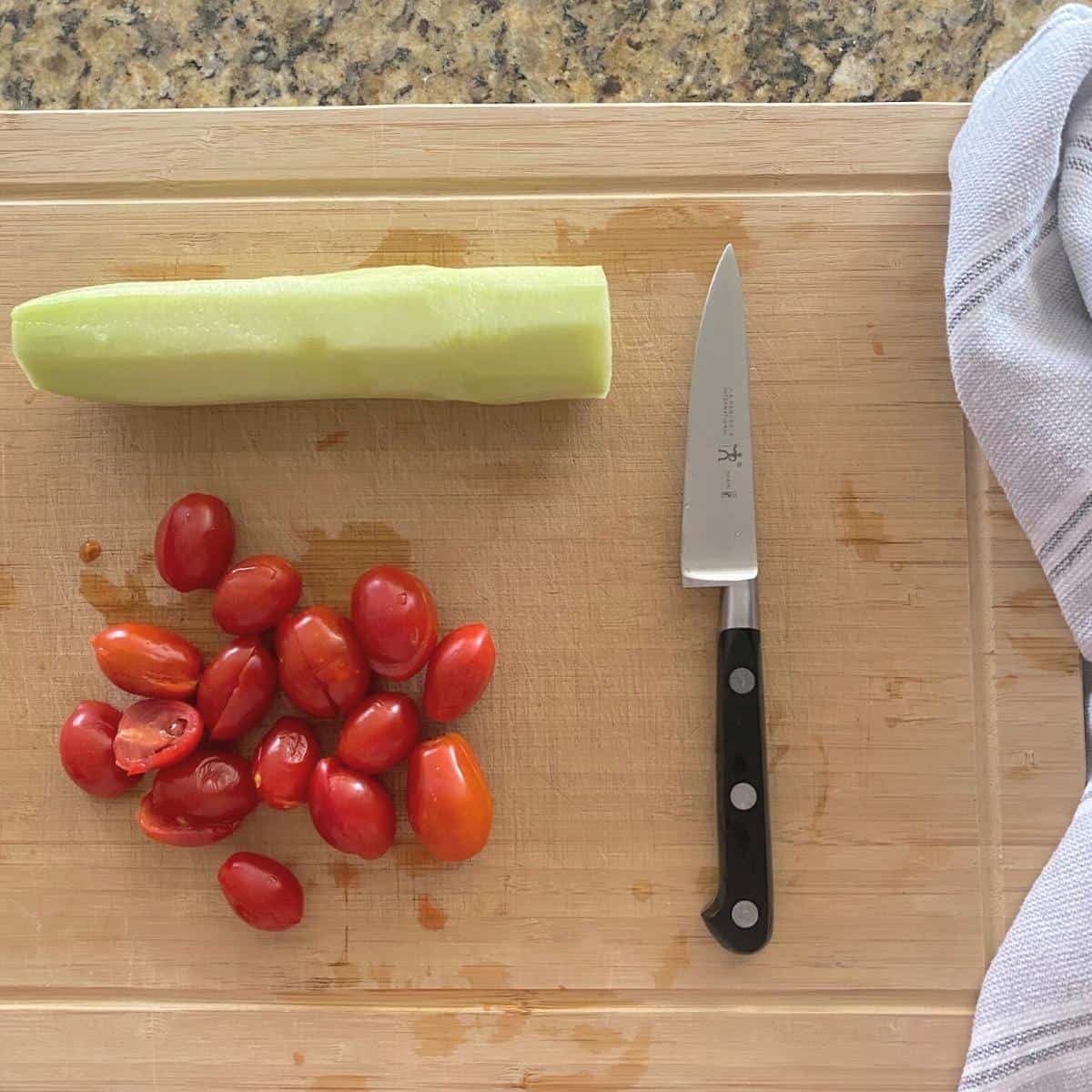 Peeled cucumber and halved grape tomatoes for salad on a cutting board next to a knife