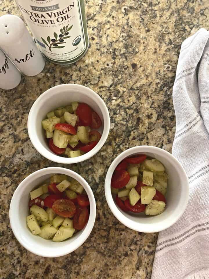 Cucumber tomato salad spread among three white ramekins, flanked atop by white salt and pepper shakers and a tin container of olive oil.