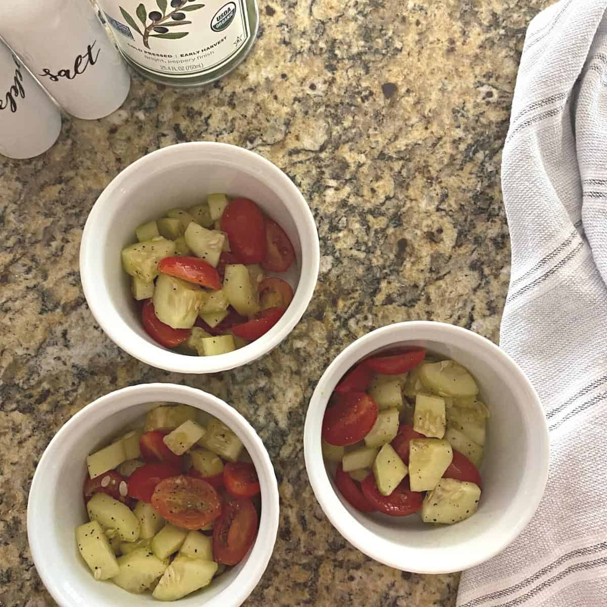 Cucumber tomato salad served in three white ramekins, flanked atop by white salt and pepper shakers and a tin container of olive oil.