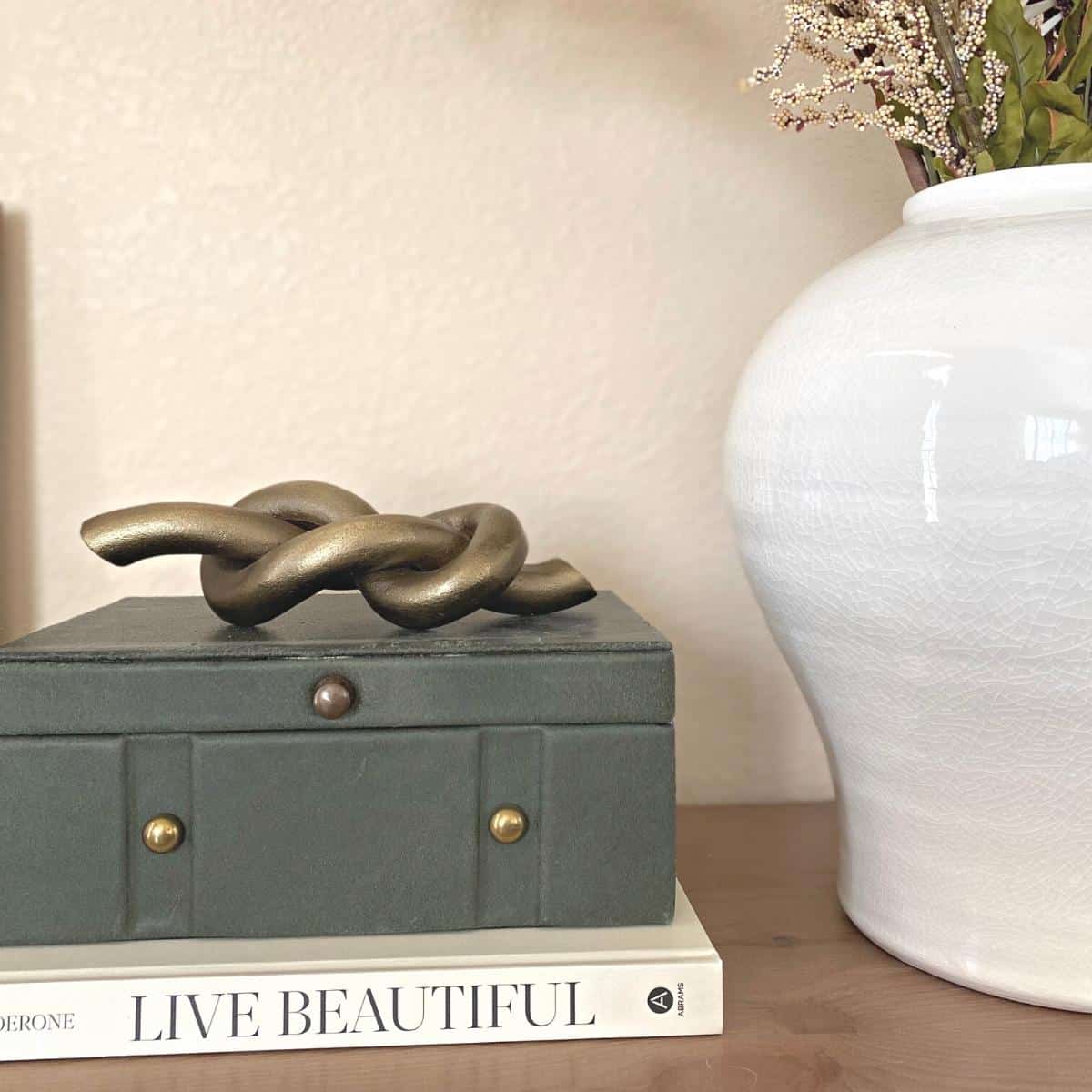 A brass knot object sits atop a dark green felt covered box with brass hardware on top of a neutral book next to a large white vase