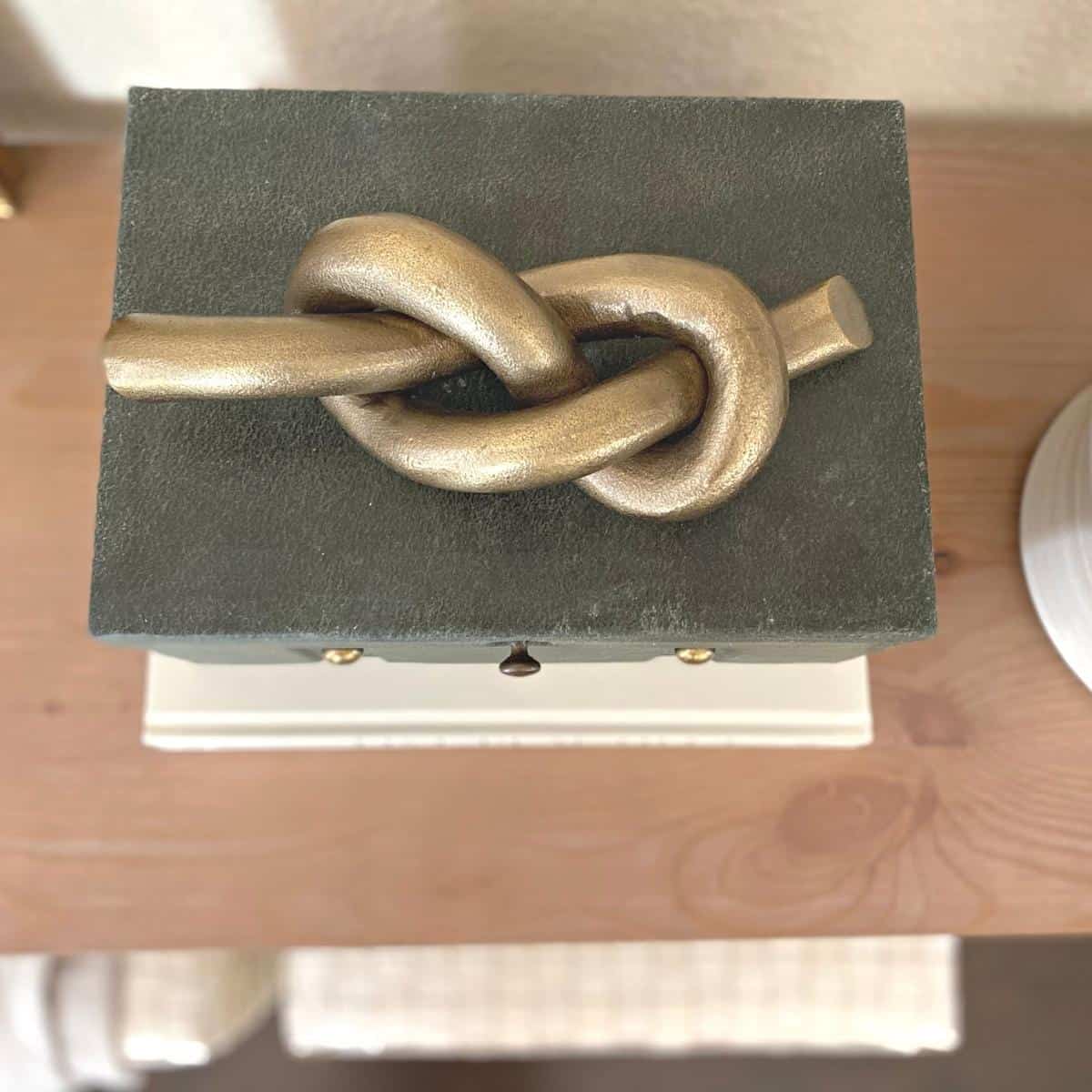 A brass knot object sits atop a dark green felt covered box with brass hardware on top of a neutral book