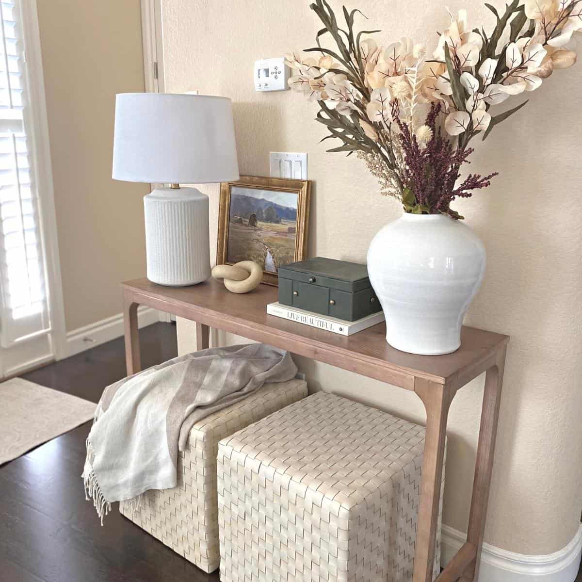 Console table decorated for fall with floral stems, a gold framed green artwork, white lamp and dark green felt covered box with brass hardware on top of a neutral book