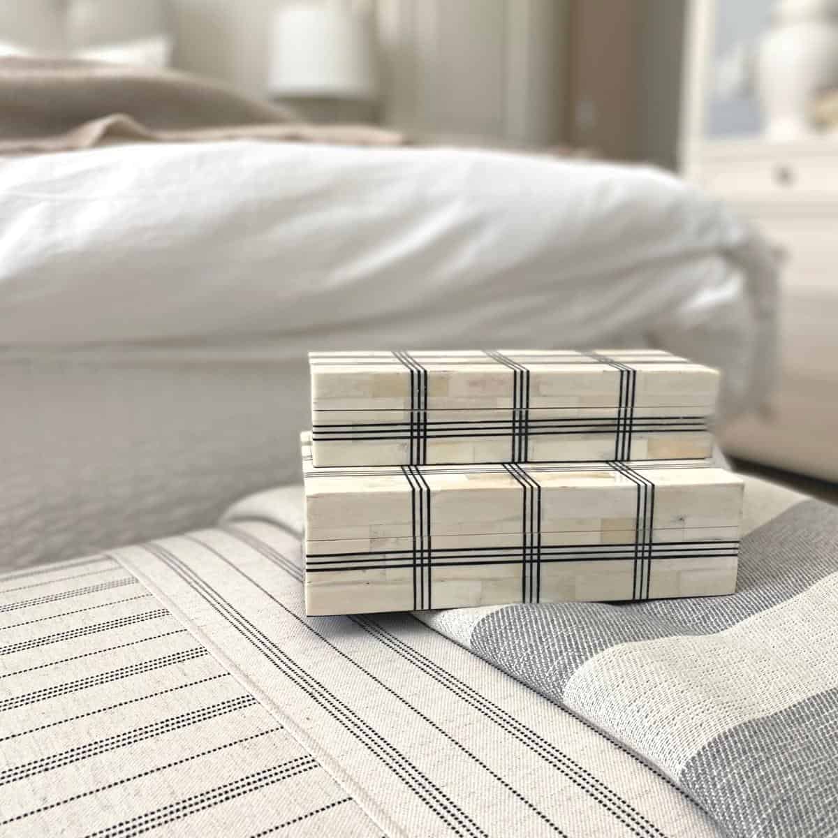 Wide grid bone boxes on top of a striped ottoman and grey-blue throw blanket at the foot of a bed