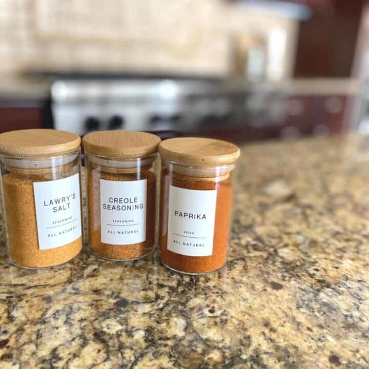 Three glass spice jars with bamboo lids and sophisticated white labels sit atop a mottled brown granite kitchen countertop. Spices are Lawry's salt, creole seasoning and paprika.