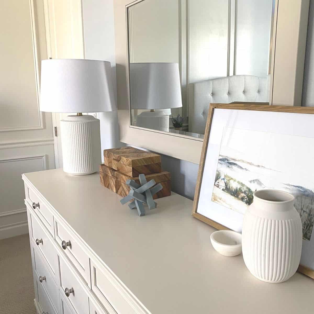 An after photo of the mirror styled hung above a white dresser with a lamp on the far left, stacked herringbone wood boxes in the middle and framed artwork on the far right, with a vase and small marble bowl in front of the artwork.