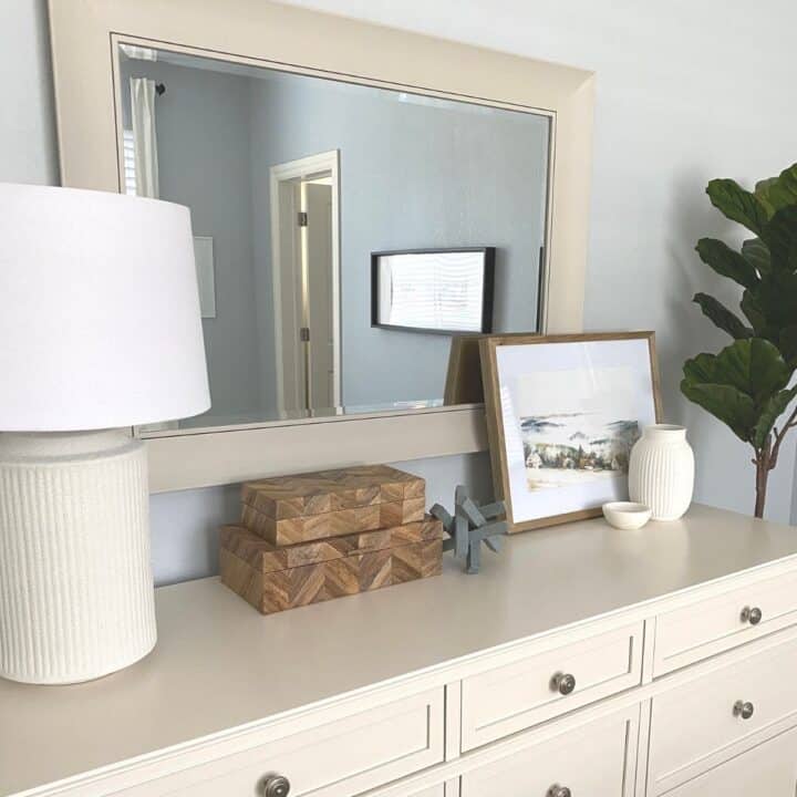 An after photo from the left side of the room showing the mirror styled hung above a white dresser with a lamp on the far left, stacked herringbone wood boxes in the middle and framed artwork on the far right, with a vase and small marble bowl in front of the artwork.