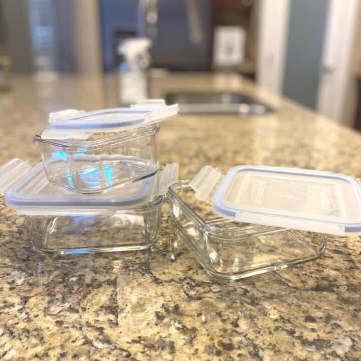 Three glass food storage containers with light blue silicone lids on a brown variegated granite countertop.