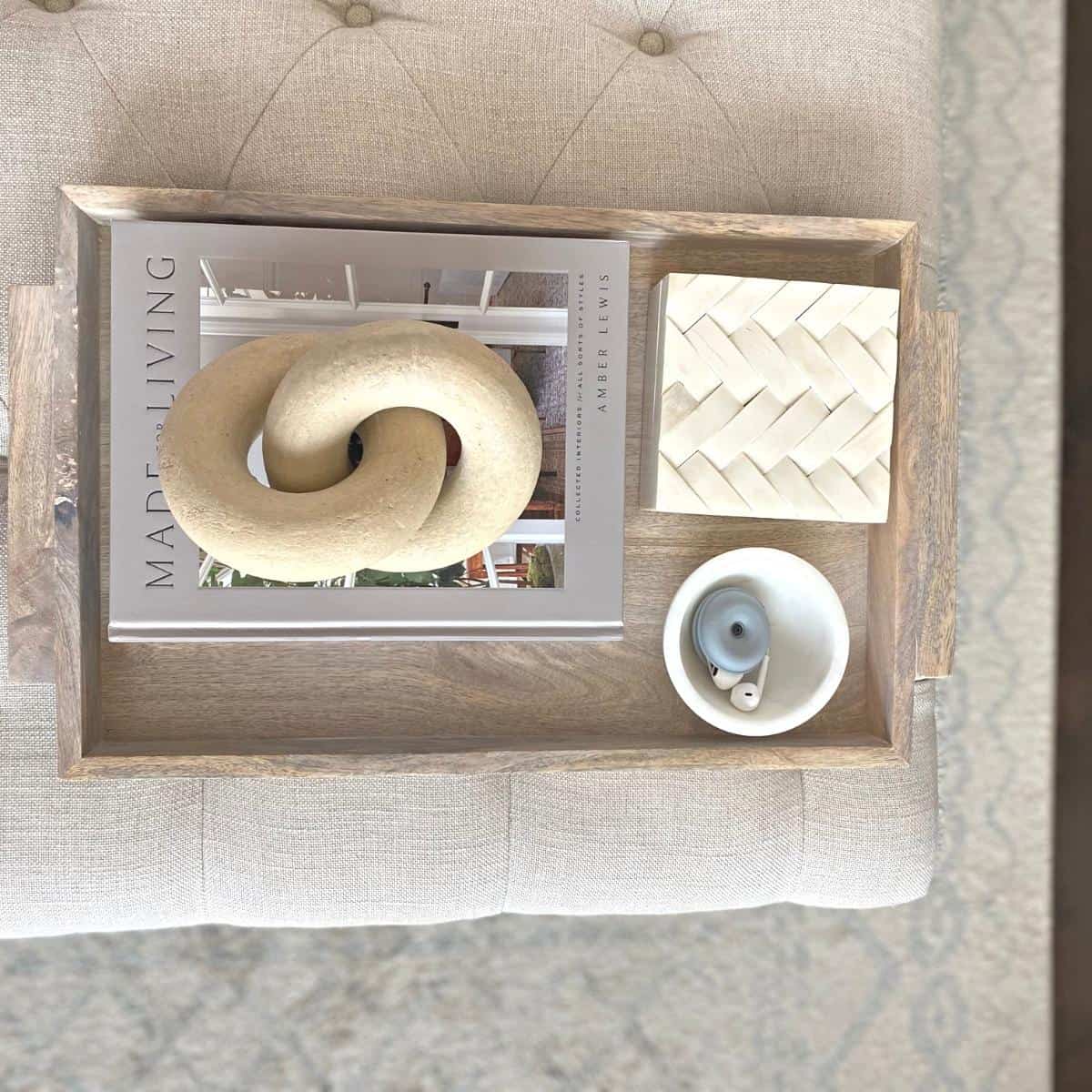 A wood tray containing a book, object, small marble bowl and bone box on an oatmeal colored tufted ottoman