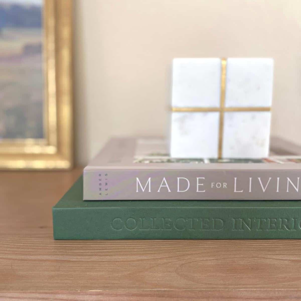 A marble and brass cube object sits atop two books; one gray and one dark green, next to gold framed artwork