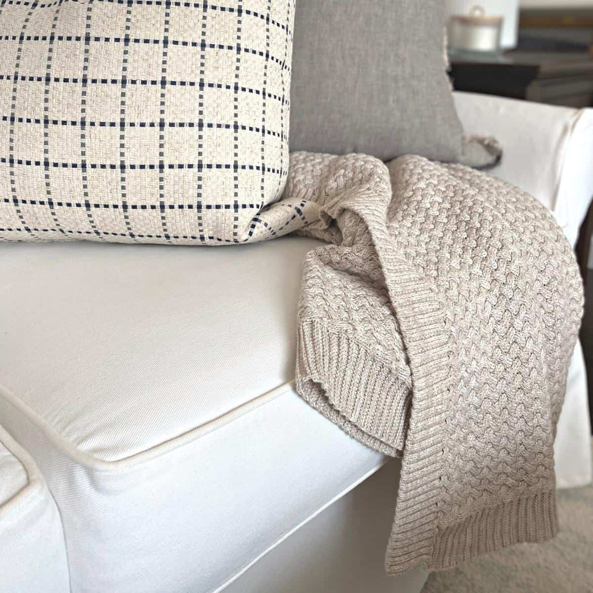 Oatmeal colored cable knit blanket draped over a white sofa with two large pillows on top