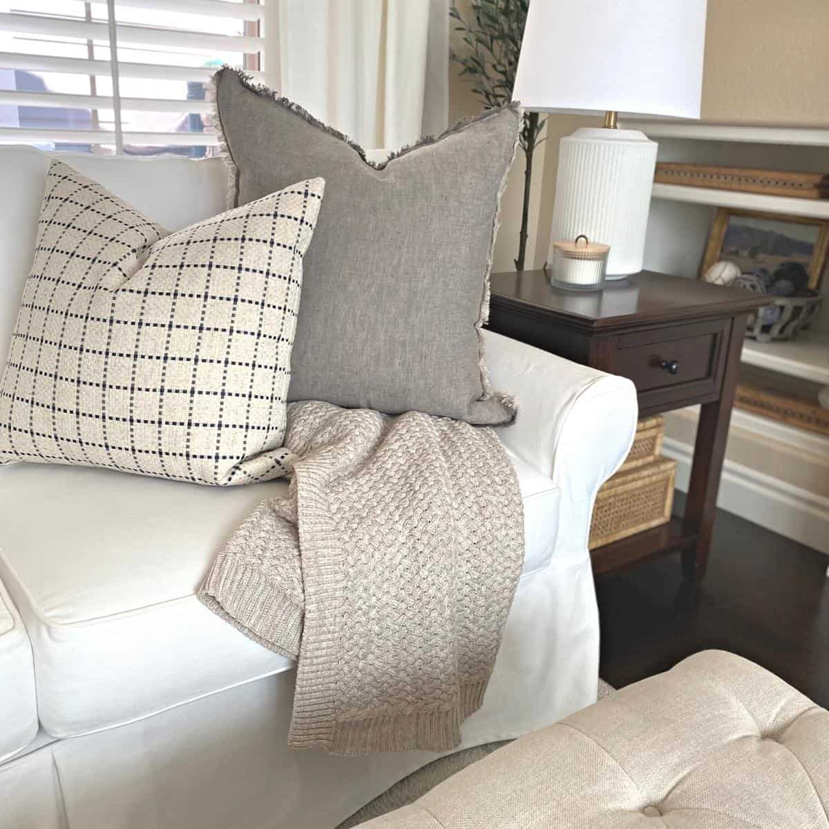 Oatmeal colored cable knit blanket draped over a white sofa with two large pillows on top