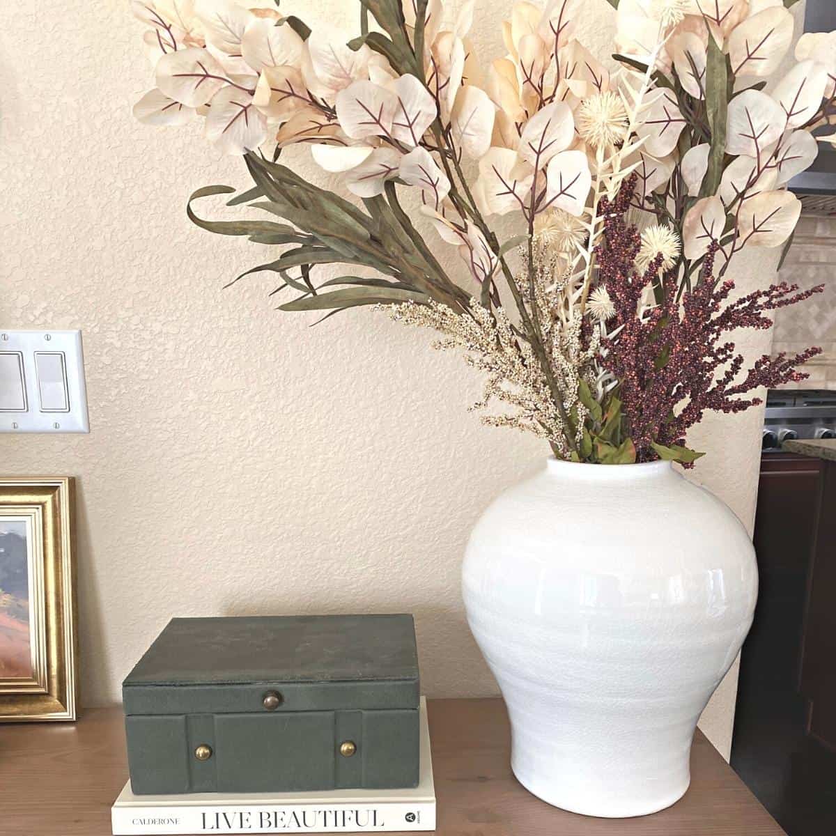 A brass knot object sits atop a dark green felt covered box with brass hardware on top of a neutral book next to a large white vase with fall floral stems