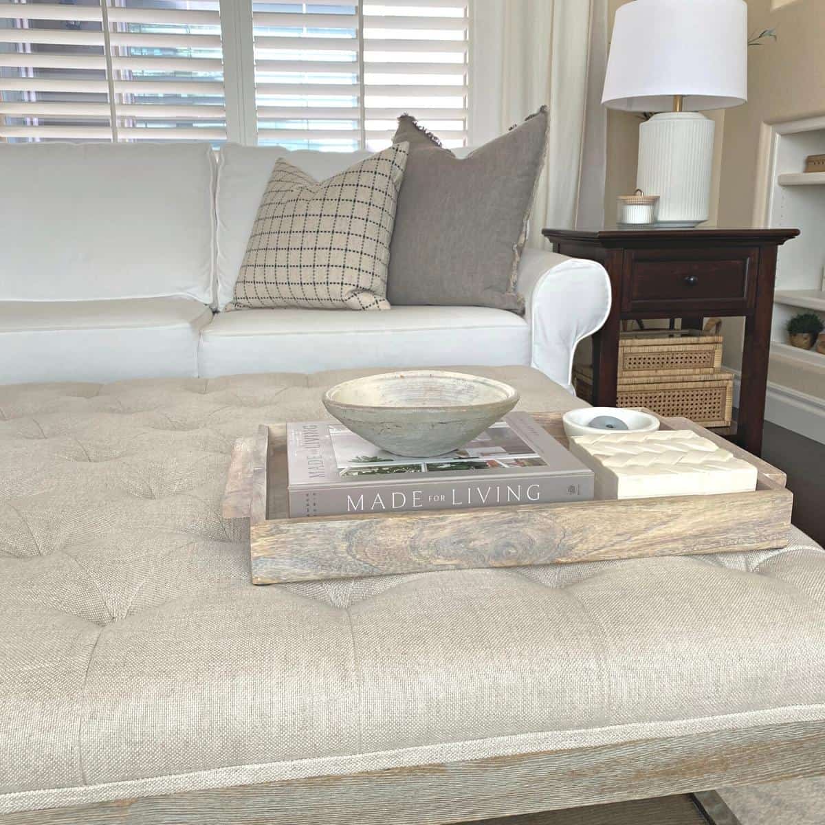 A wood tray containing a book, object, small marble bowl and bone box on an oatmeal colored tufted ottoman