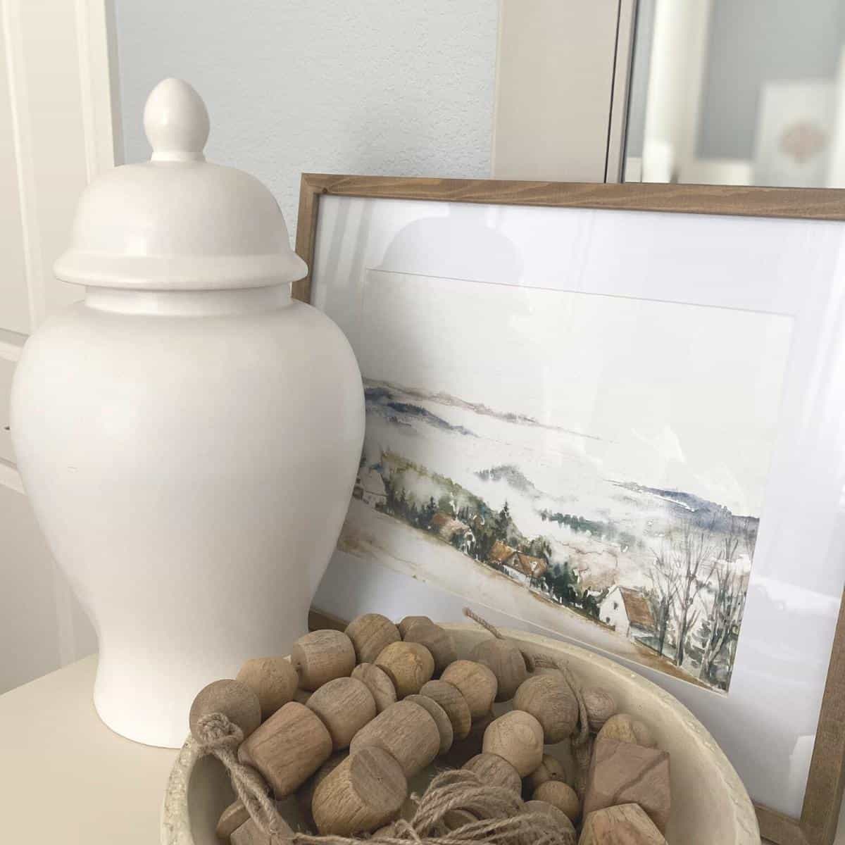 Framed mountainside artwork styled on a dresser, with large white ginger jar and bowl of wood bead garland