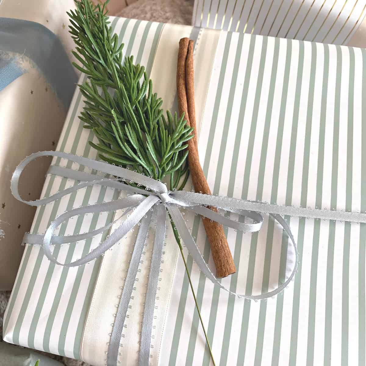 A present wrapped in a gray-green striped paper with a sprig of greenery, a cinnamon stick and a blue satin ribbon bow