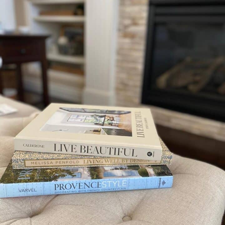 Coffee table books stacked on a living room flax color linen fabric covered ottoman. The three books are Living Well by Design by Melissa Penfold, Provence Style colon Decorating with French Country Flair by Shauna Varvel and Live Beautiful by Athena Calderone.