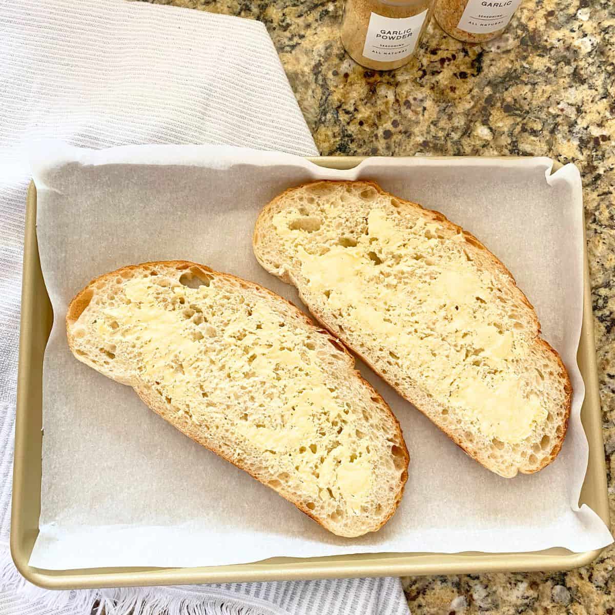 Buttered and garlic powdered sourdough bread on a gold baking sheet pan, waiting to be roasted in the oven
