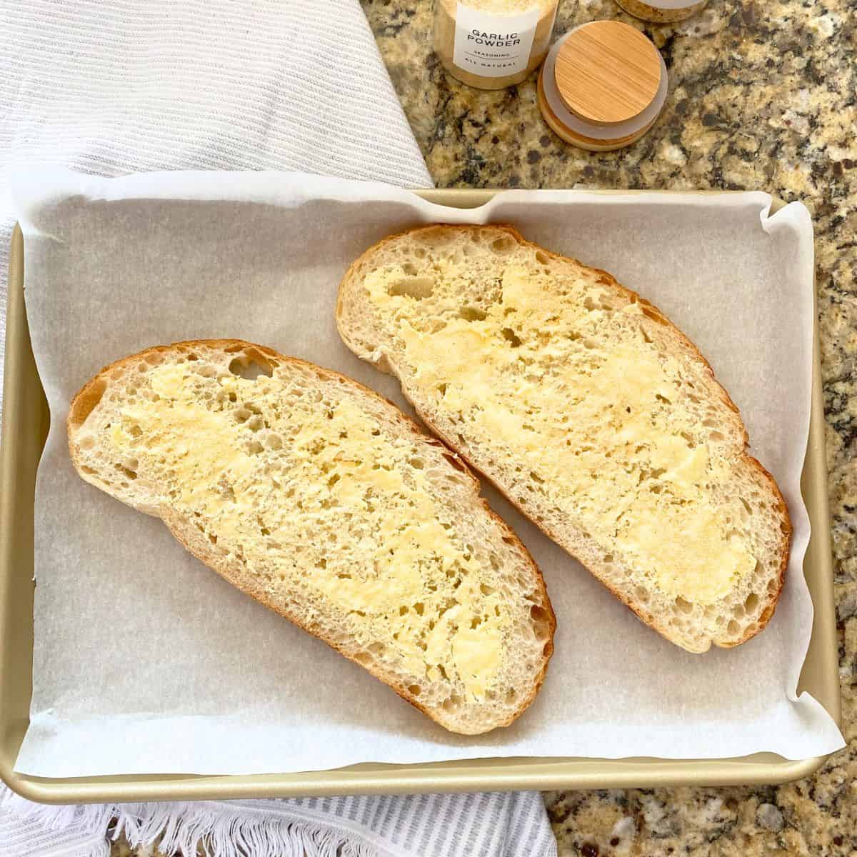 Buttered sourdough bread on a gold baking sheet pan, waiting to be roasted in the oven