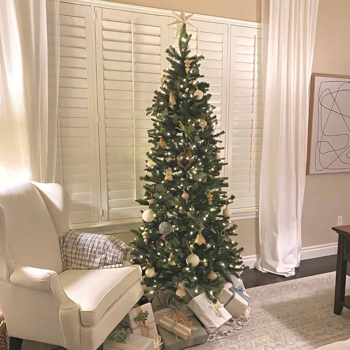 A decorated pre-lit Christmas tree with neutral ornaments placed in the front window of a living room