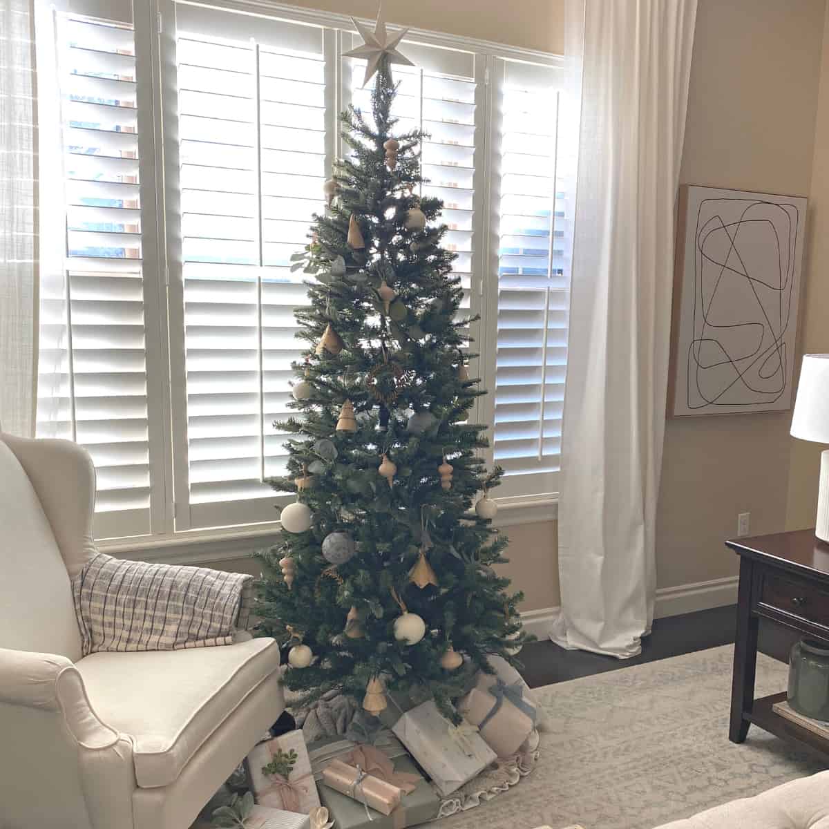 A decorated Christmas tree with neutral ornaments and presents beneath is placed in the front window of a living room