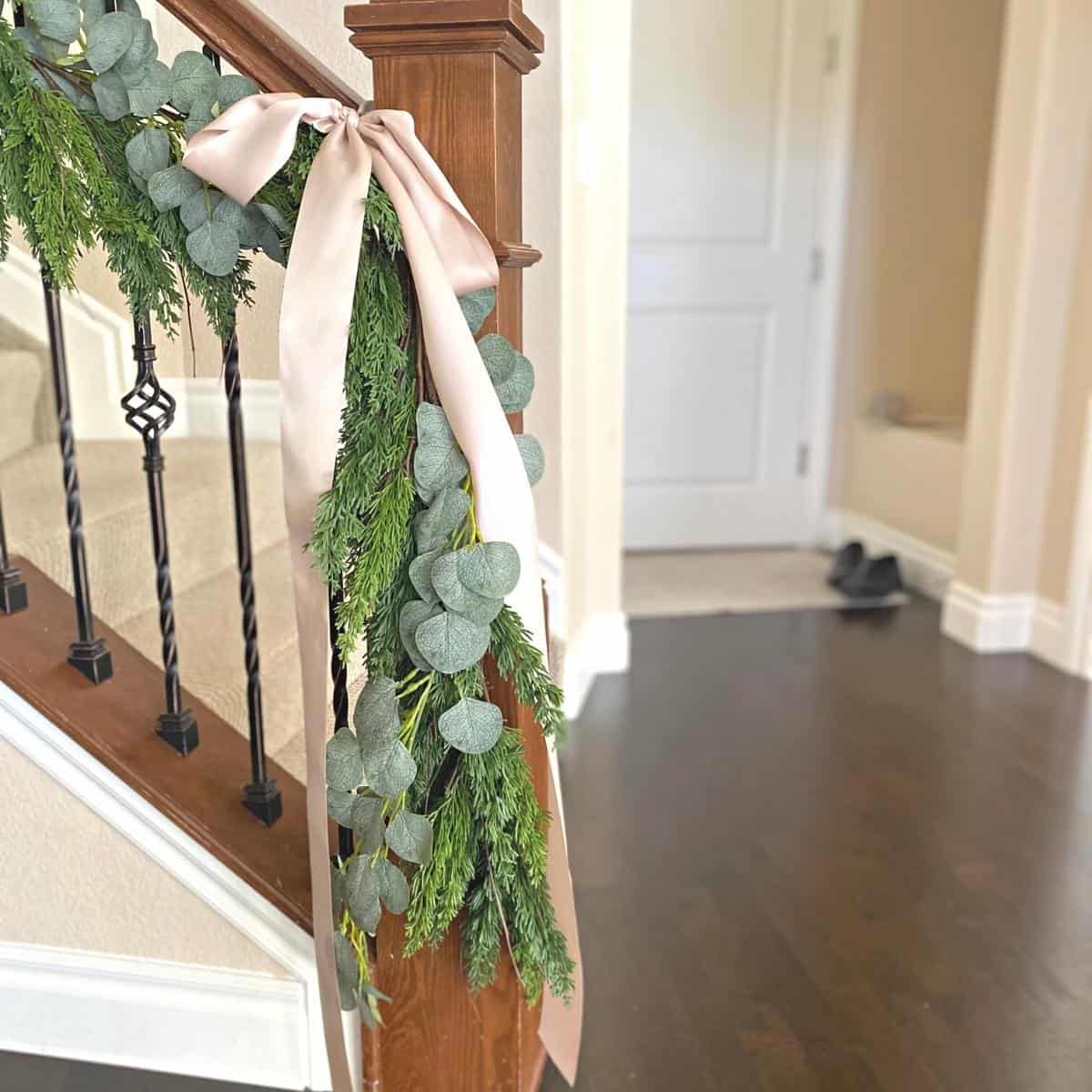 Taupe-pink ribbon bows hanging on staircase handrail holiday garland of faux cedar and eucalyptus