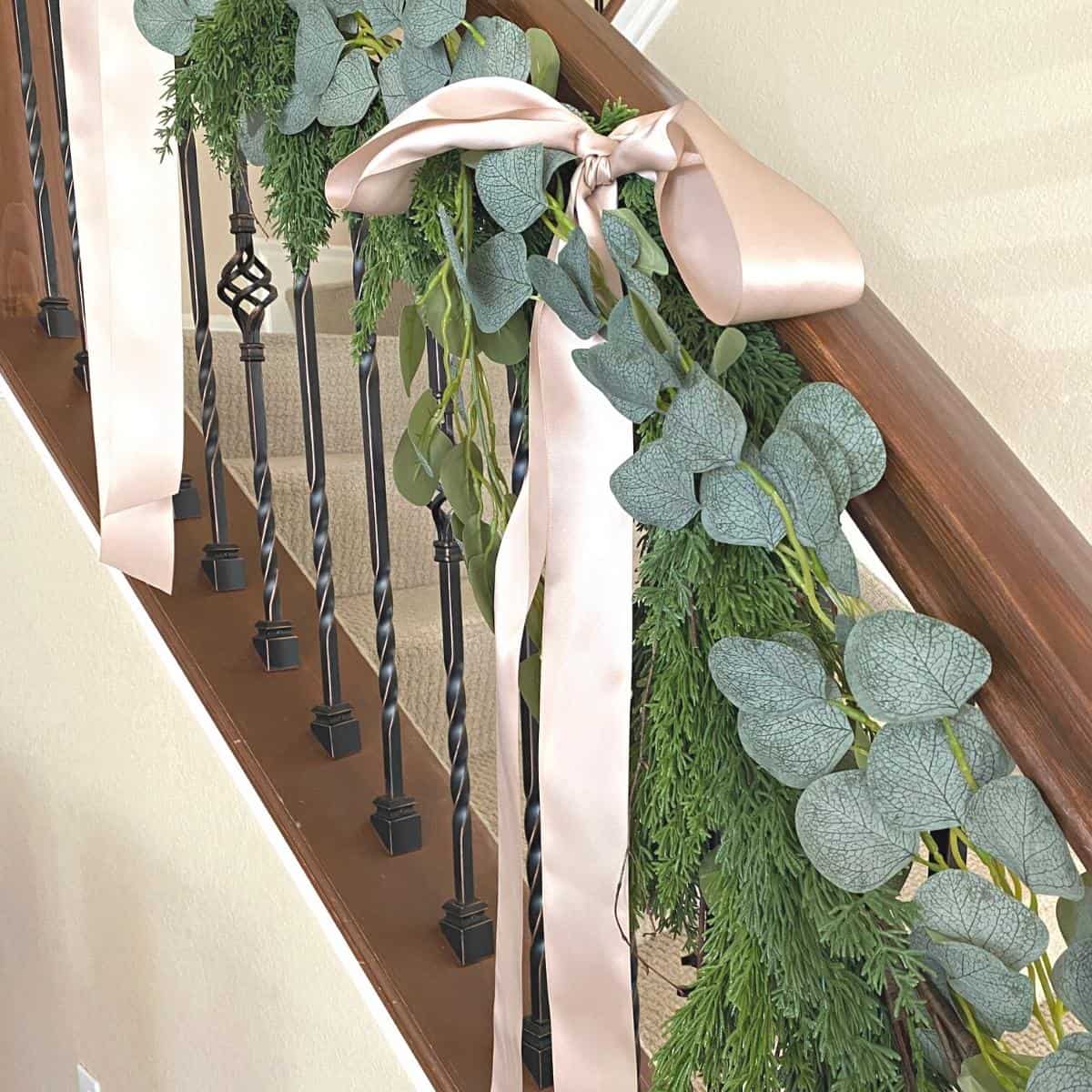 Taupe-pink ribbon bows hanging on staircase handrail holiday garland of faux cedar and eucalyptus