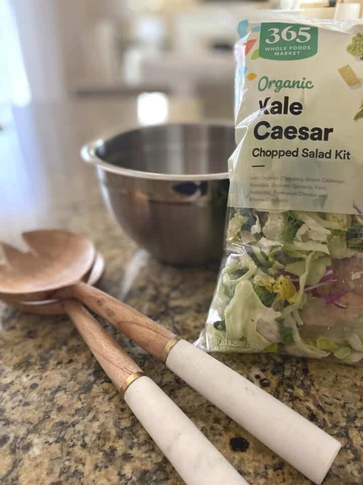 Kale Caesar salad flanks a stylish wood and marble salad server set and a small stainless steel mixing bowl.