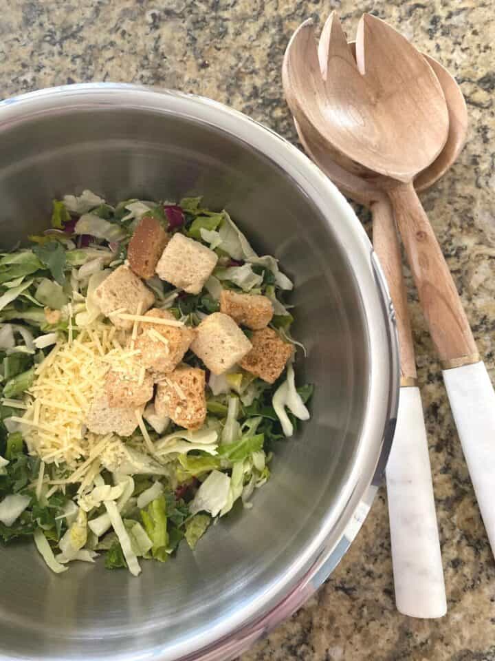 Kale Caesar salad with toppings in a small stainless steel mixing bowl next to a stylish wood and marble salad server set