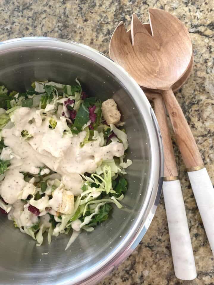 Kale Caesar salad with toppings and dressing in a small stainless steel mixing bowl next to a stylish wood and marble salad server set